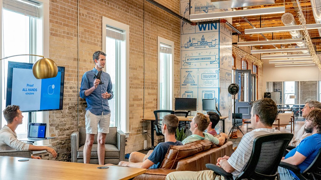 White man standing in front of a screen delivering a presentation to a male audience.