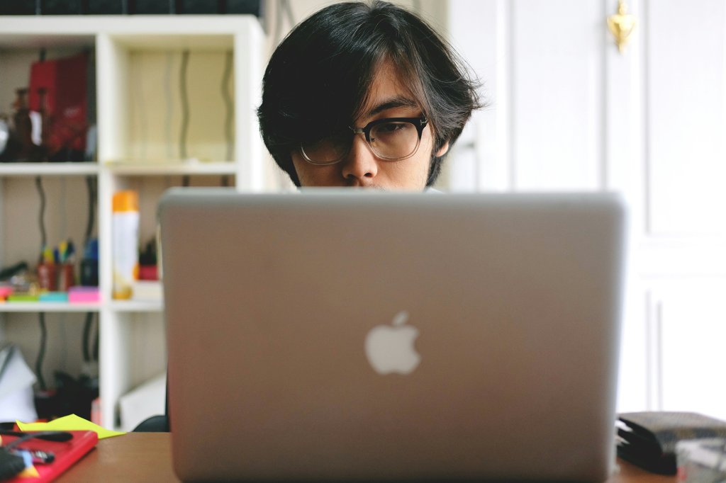 Young Asian man in thick glasses sitting behind a MacBook laptop.